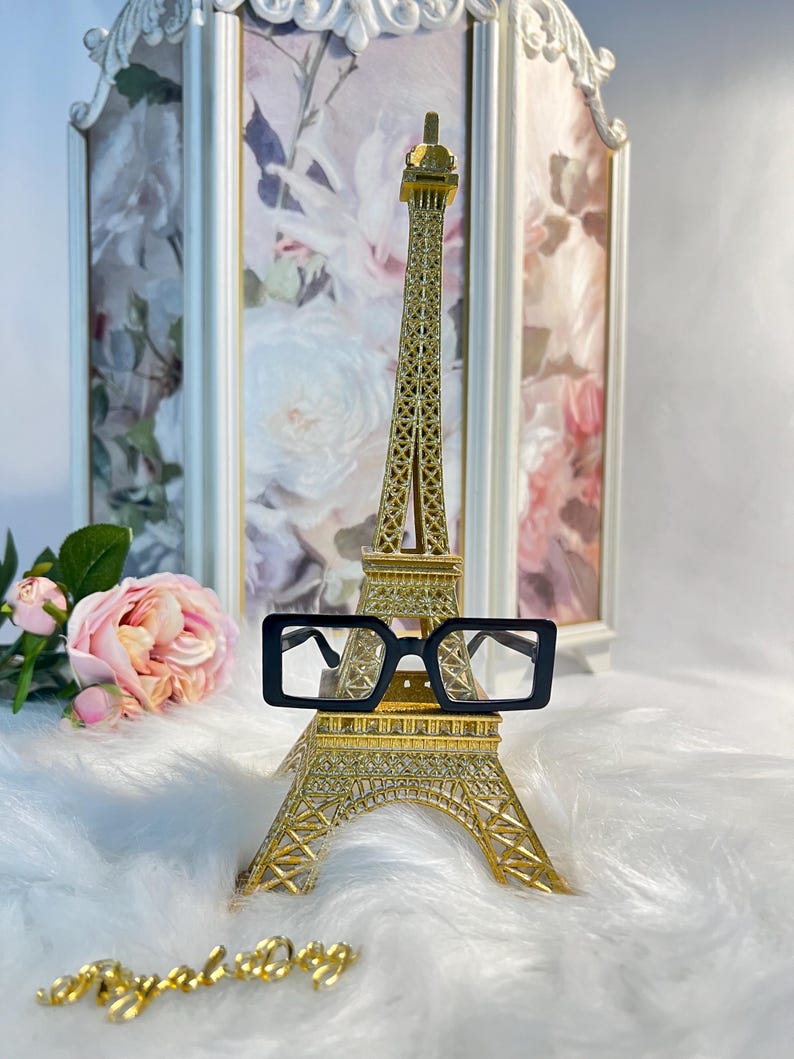 May include: Black square-shaped eyeglasses with a gold Eiffel Tower miniature in the background. The Eiffel Tower is on a white fluffy surface.