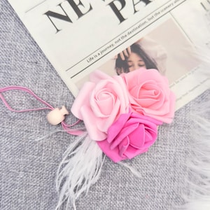 May include: A floral accessory featuring three fabric roses in shades of pink, accented with white feathers and a small wooden bead. The roses rest on a newspaper with the headline "THE NEWSPAPER PARIS".