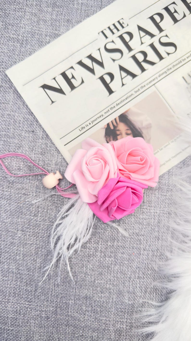 May include: A floral accessory featuring three fabric roses in shades of pink, accented with white feathers and a small wooden bead. The roses rest on a newspaper with the headline "THE NEWSPAPER PARIS".