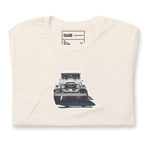 May include: Cream-colored t-shirt featuring a graphic of a white vintage Toyota Land Cruiser. The shirt has a crew neck and short sleeves. The text "five CAR GARAGE - Adam Ambro" is printed on the tag.