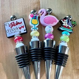 May include: Four decorative wine bottle stoppers with unique designs. One features the text "It's Wine O'Clock Somewhere" and wine glasses. Other stoppers have a cocktail, a pink flamingo, and a Santa Claus on a boat. Each has a silver base.
