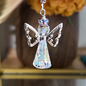 May include: A silver angel pendant necklace with a clear, iridescent crystal body and wings. The angel has a halo of iridescent crystals and hangs from a delicate silver chain. The background is blurred, with a vase of flowers.