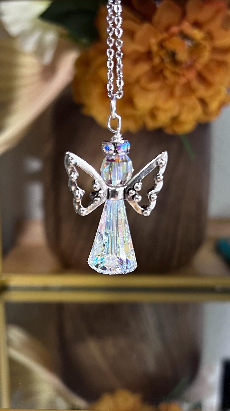 May include: A silver angel pendant necklace with a clear, iridescent crystal body and wings. The angel has a halo of iridescent crystals and hangs from a delicate silver chain. The background is blurred, with a vase of flowers.