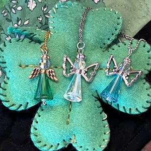 May include: Three angel pendant necklaces with clear, green, and blue crystal bodies and silver or gold-tone wings. The pendants hang on delicate chains and rest on a green felt shamrock.