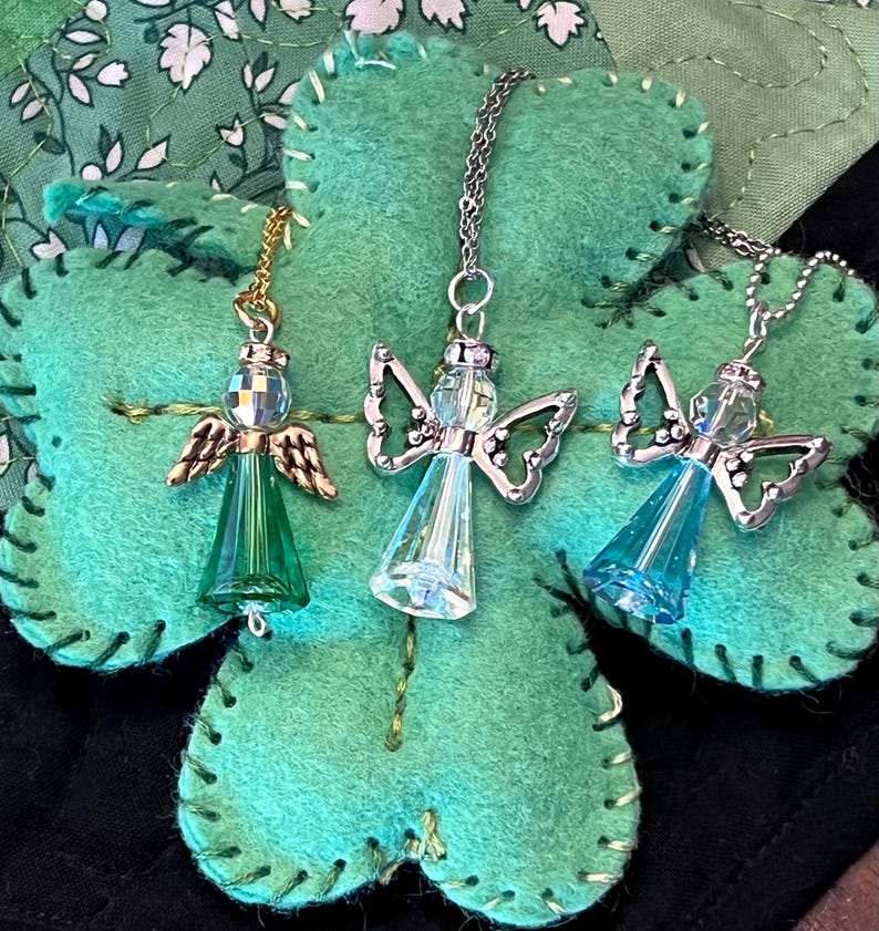May include: Three angel pendant necklaces with clear, green, and blue crystal bodies and silver or gold-tone wings. The pendants hang on delicate chains and rest on a green felt shamrock.