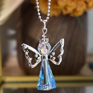 May include: A silver-toned angel pendant necklace. The angel has a blue crystal body, iridescent head, and detailed wings. The necklace chain is a delicate ball chain, perfect for everyday wear.