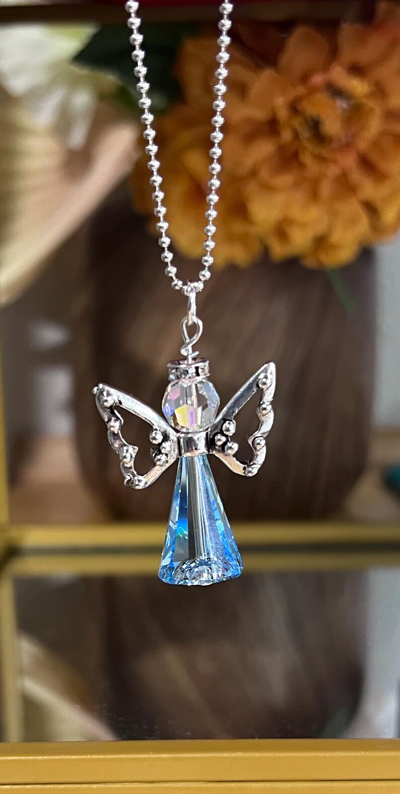May include: A silver-toned angel pendant necklace. The angel has a blue crystal body, iridescent head, and detailed wings. The necklace chain is a delicate ball chain, perfect for everyday wear.
