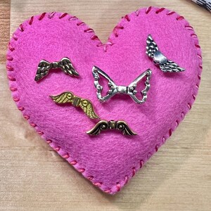 May include: A pink felt heart with red stitching, adorned with five metal wing-shaped embellishments in silver and gold tones. The heart shape is prominent, and the wings are detailed.