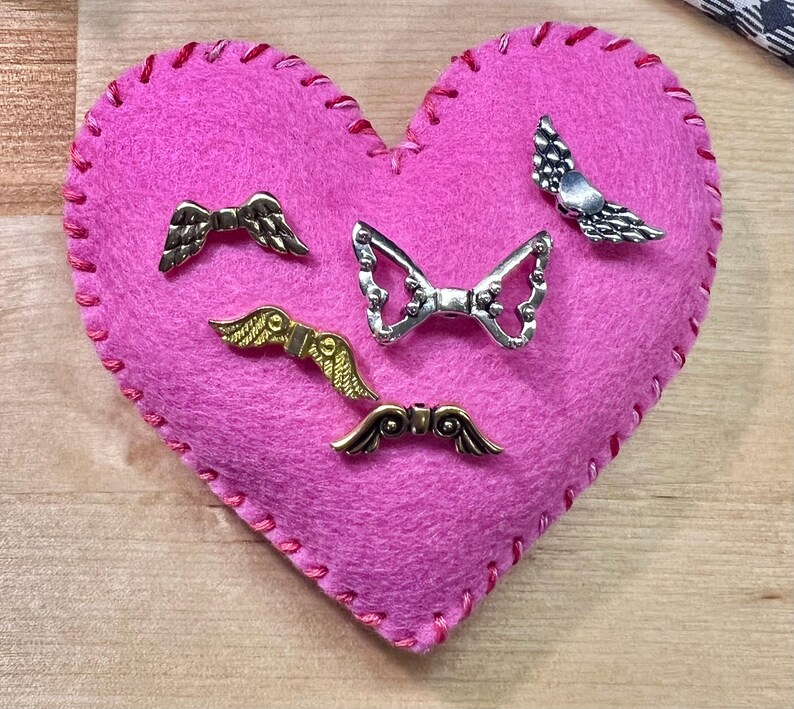 May include: A pink felt heart with red stitching, adorned with five metal wing-shaped embellishments in silver and gold tones. The heart shape is prominent, and the wings are detailed.