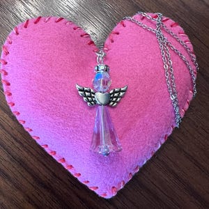 May include: A silver angel pendant necklace with a clear crystal body and wings, resting on a pink felt heart with red stitching. The necklace chain is silver. The background is a dark wood surface.