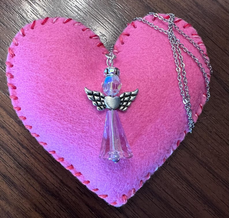 May include: A silver angel pendant necklace with a clear crystal body and wings, resting on a pink felt heart with red stitching. The necklace chain is silver. The background is a dark wood surface.