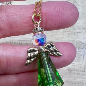 May include: A gold-colored chain necklace with a pendant in the shape of an angel. The angel pendant features a faceted, iridescent crystal head, gold-colored wings, and a green, faceted body. The necklace is held by a person's fingers.