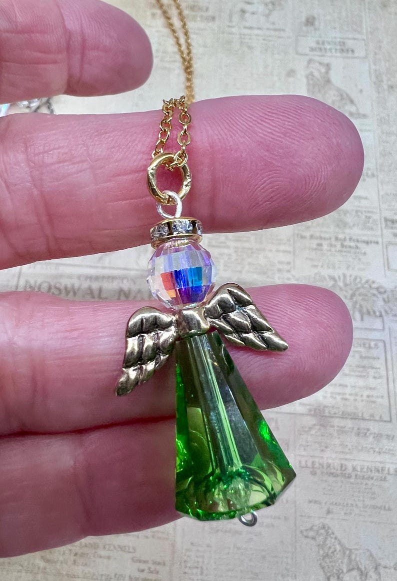 May include: A gold-colored chain necklace with a pendant in the shape of an angel. The angel pendant features a faceted, iridescent crystal head, gold-colored wings, and a green, faceted body. The necklace is held by a person's fingers.