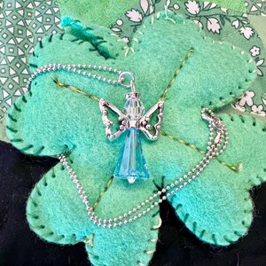 May include: A silver angel pendant with a blue crystal body and detailed wings, suspended on a delicate silver chain. The necklace rests on a large, light green felt shamrock with stitched edges, set against a patchwork background.