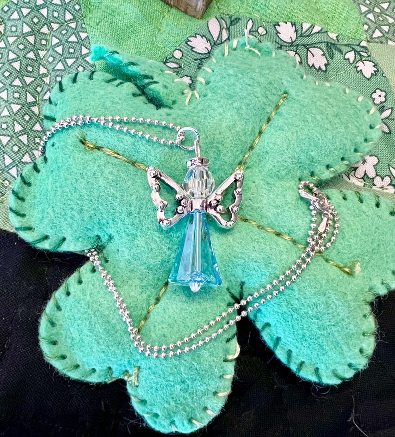 May include: A silver angel pendant with a blue crystal body and detailed wings, suspended on a delicate silver chain. The necklace rests on a large, light green felt shamrock with stitched edges, set against a patchwork background.