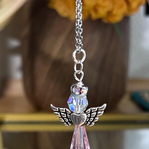 May include: A silver-toned angel pendant with a chain. The angel features a crystal head, heart-shaped body, and wings. The lower portion is a faceted, light purple crystal. The background includes a blurred orange flower and a gold-framed display case.