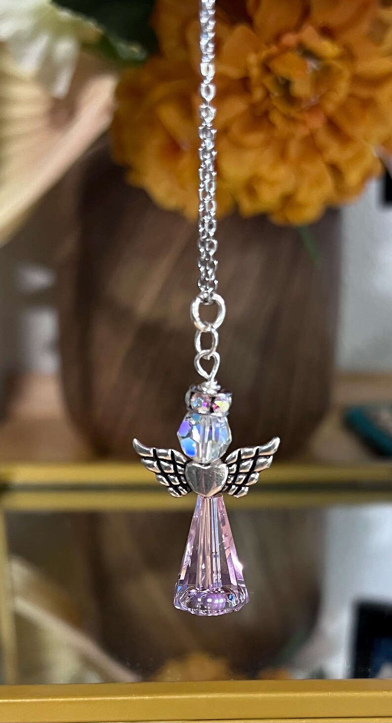 May include: A silver-toned angel pendant with a chain. The angel features a crystal head, heart-shaped body, and wings. The lower portion is a faceted, light purple crystal. The background includes a blurred orange flower and a gold-framed display case.