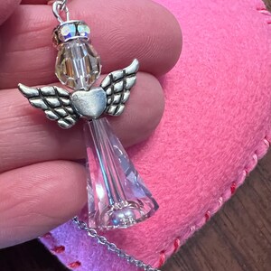 May include: A silver angel pendant with crystal wings and a heart-shaped body. The angel is suspended from a delicate chain. A pink felt heart with red stitching is in the background.