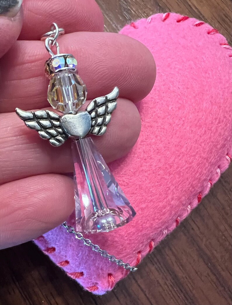 May include: A silver angel pendant with crystal wings and a heart-shaped body. The angel is suspended from a delicate chain. A pink felt heart with red stitching is in the background.