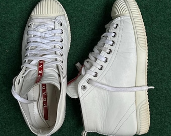 May include: Pair of white high-top trainers with white laces and a ridged rubber sole. The trainers have a red tag with the brand name "PRADA". The shoes are on a green surface.