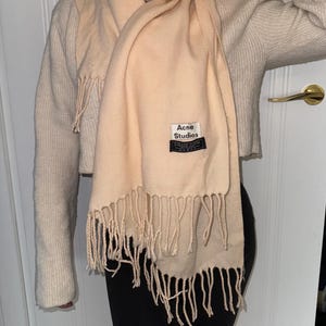 May include: A pale pink scarf with fringe detailing and a black label that reads "Acne Studios" in white lettering.