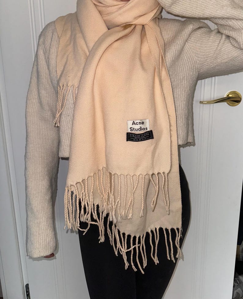 May include: A pale pink scarf with fringe detailing and a black label that reads "Acne Studios" in white lettering.