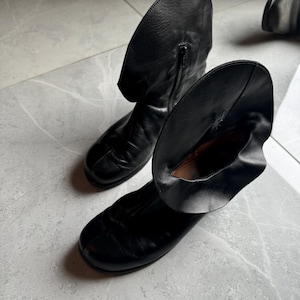 May include: A pair of black leather boots with a unique design. The boots have a rounded toe and a folded-over cuff. A zipper is visible on the side of one boot. The boots are on a light gray surface.