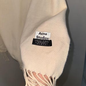 May include: A cream-coloured wool scarf with fringe. The scarf has a black label that reads "Acne Studios" and "Virgin Wool" with details about the material and origin.