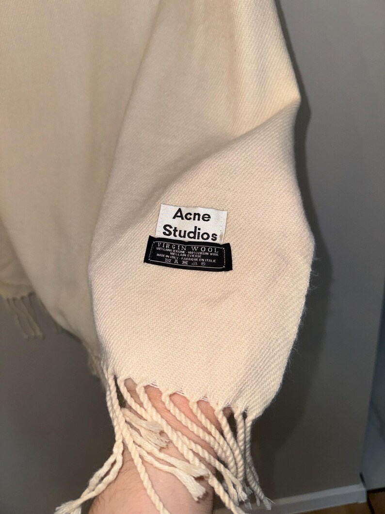 May include: A cream-coloured wool scarf with fringe. The scarf has a black label that reads "Acne Studios" and "Virgin Wool" with details about the material and origin.