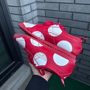Puede incluir: Un par de zapatillas rojas con dise&ntilde;os circulares y geom&eacute;tricos blancos. Los zapatos tienen cordones y suelas rojas. Las zapatillas se sostienen contra una pared de ladrillos grises.