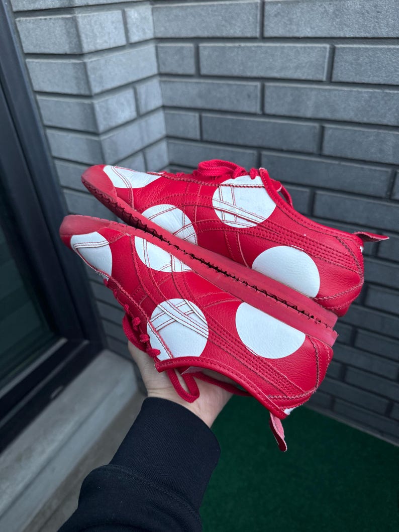 Puede incluir: Un par de zapatillas rojas con dise&ntilde;os circulares y geom&eacute;tricos blancos. Los zapatos tienen cordones y suelas rojas. Las zapatillas se sostienen contra una pared de ladrillos grises.