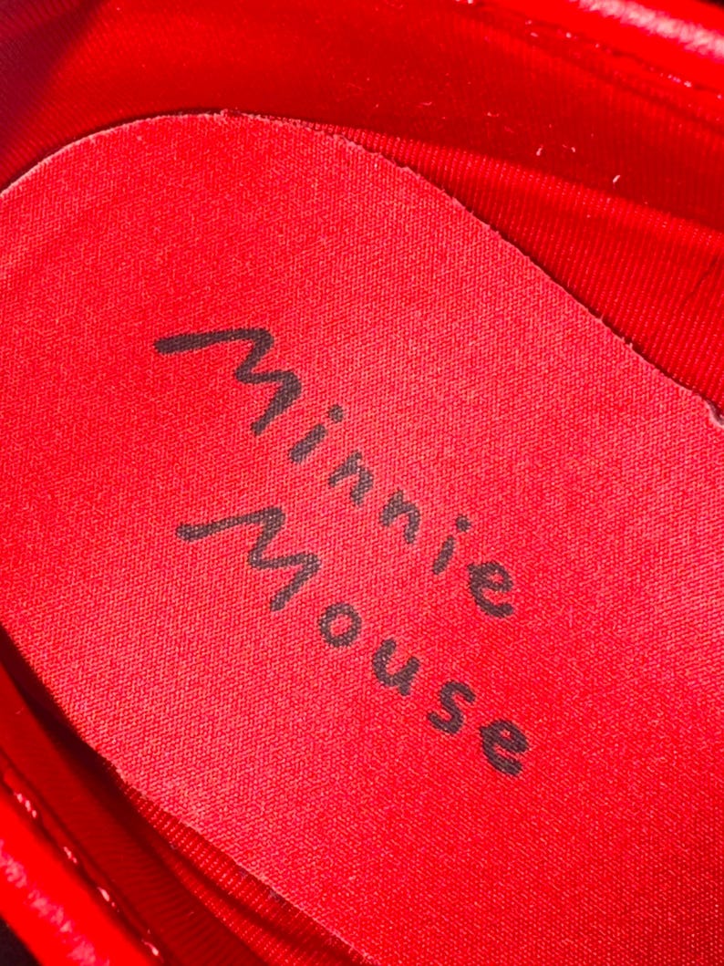 Puede incluir: Primer plano del interior de un zapato rojo. La plantilla es roja con las palabras "Minnie Mouse" impresas en escritura cursiva negra. El interior del zapato tambi&eacute;n es rojo.