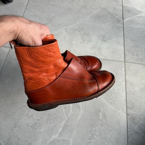 May include: A pair of ankle-high leather boots in shades of orange and brown. The boots have a textured orange upper and a smooth brown lower section. The soles are dark brown with visible stitching. The boots are held by a hand.
