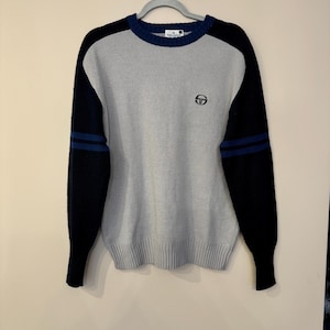 Puede incluir: Un suéter de manga larga gris y negro con rayas azules en las mangas. El suéter tiene cuello redondo y un pequeño logotipo en la parte delantera.