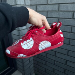 Puede incluir: Zapatillas rojas con lunares blancos y rayas laterales blancas. Los zapatos tienen cordones rojos y una suela roja. El tal&oacute;n presenta un dise&ntilde;o blanco. Las zapatillas son sostenidas por una persona.