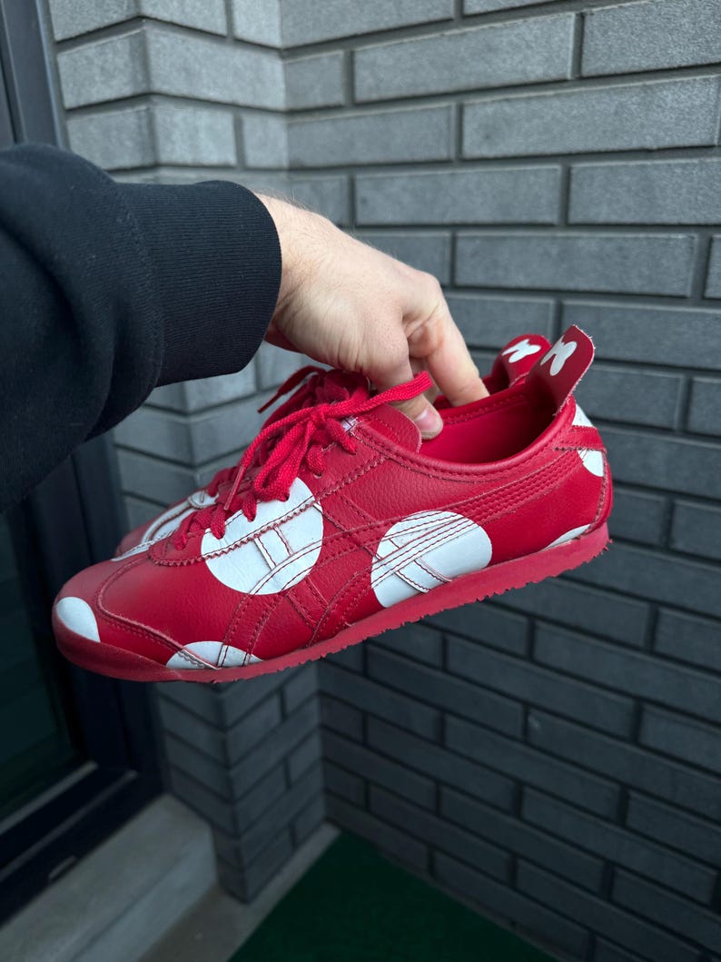 Puede incluir: Zapatillas rojas con lunares blancos y rayas laterales blancas. Los zapatos tienen cordones rojos y una suela roja. El tal&oacute;n presenta un dise&ntilde;o blanco. Las zapatillas son sostenidas por una persona.