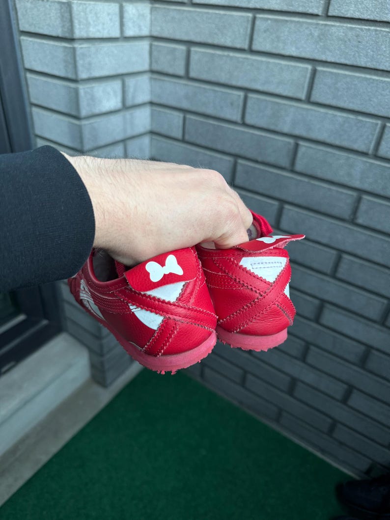 Puede incluir: Un par de zapatillas rojas con detalles blancos y un lazo en el tal&oacute;n. Las zapatillas tienen un perfil bajo y una suela de goma. Las zapatillas son sostenidas por una persona.