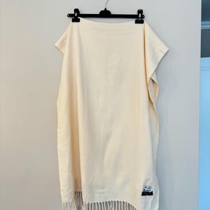 May include: A cream-coloured woven scarf with fringe on the bottom edge. The scarf has a small black and white label that says "Acne Studios" on it.