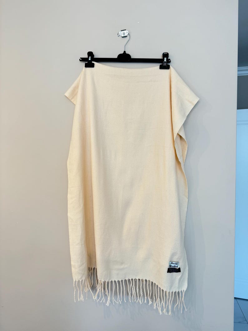 May include: A cream-coloured woven scarf with fringe on the bottom edge. The scarf has a small black and white label that says "Acne Studios" on it.