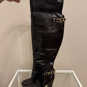 Puede incluir: Botas negras de cuero por encima de la rodilla con estampado de cocodrilo y detalles de cadena dorada. Las botas tienen un tacón alto.