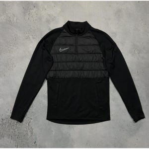 Könnte beinhalten: Schwarzes Langarm-Shirt mit 1/4-Reißverschluss und einem gesteppten Einsatz auf der Brust. Das Shirt hat ein weißes Nike-Logo auf der Brust.