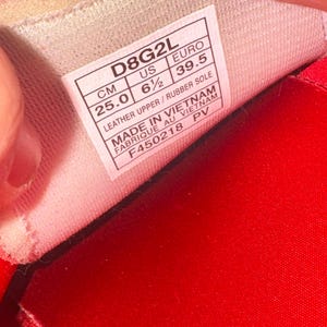 Puede incluir: Primer plano del interior de un zapato, con una etiqueta blanca y texto negro. La etiqueta indica una talla de 39,5 y una medida de 25,0 cm. El zapato es rojo.
