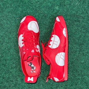 Puede incluir: Un par de zapatillas rojas con lunares blancos y cordones rojos. Los zapatos tienen un dise&ntilde;o blanco y rojo en los laterales y un gr&aacute;fico de Minnie Mouse en la plantilla. Los zapatos est&aacute;n sobre una superficie verde.