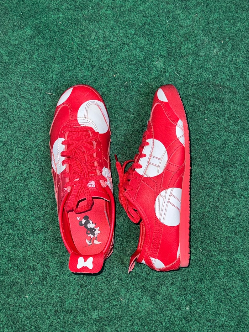 Puede incluir: Un par de zapatillas rojas con lunares blancos y cordones rojos. Los zapatos tienen un dise&ntilde;o blanco y rojo en los laterales y un gr&aacute;fico de Minnie Mouse en la plantilla. Los zapatos est&aacute;n sobre una superficie verde.