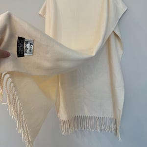 May include: A cream-coloured scarf with fringe on the ends. The scarf has a black label that reads "Acne Studios" with the text "Made in Italy" underneath.