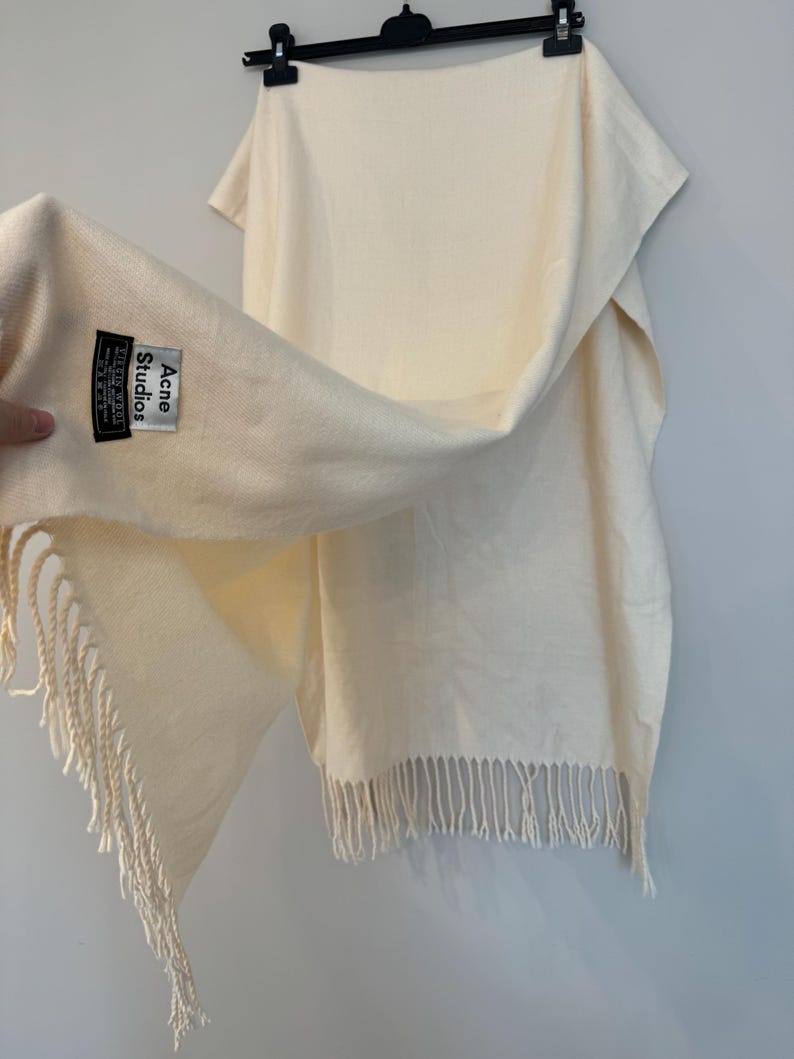 May include: A cream-coloured scarf with fringe on the ends. The scarf has a black label that reads "Acne Studios" with the text "Made in Italy" underneath.