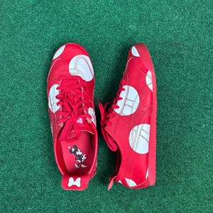 Puede incluir: Zapatillas rojas con lunares blancos y cordones rojos. Los zapatos tienen un dise&ntilde;o de rayas blancas y un gr&aacute;fico de Minnie Mouse en la plantilla. Los zapatos est&aacute;n sobre una superficie verde.