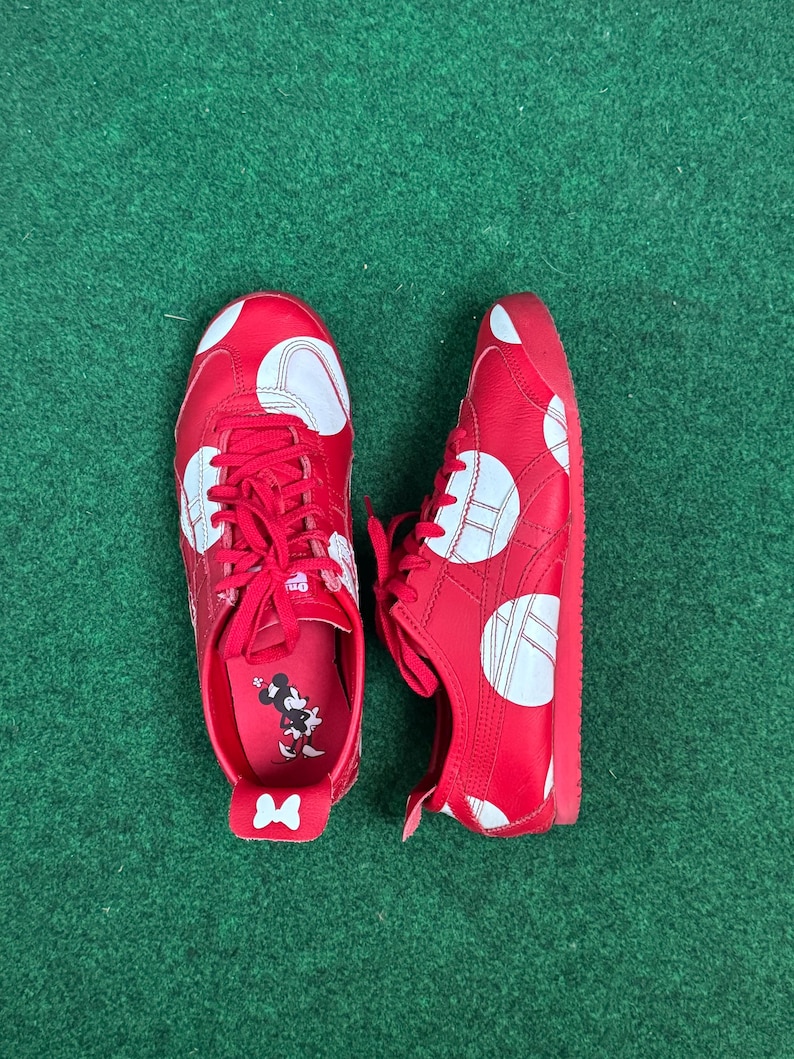 Puede incluir: Zapatillas rojas con lunares blancos y cordones rojos. Los zapatos tienen un dise&ntilde;o de rayas blancas y un gr&aacute;fico de Minnie Mouse en la plantilla. Los zapatos est&aacute;n sobre una superficie verde.