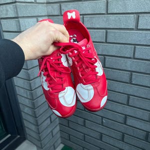 Puede incluir: Un par de zapatillas rojas con lunares y detalles blancos. Los zapatos tienen cordones rojos y un detalle de lazo blanco en la leng&uuml;eta. Las zapatillas se sostienen contra una pared de ladrillos gris.