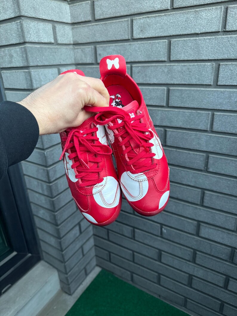 Puede incluir: Un par de zapatillas rojas con lunares y detalles blancos. Los zapatos tienen cordones rojos y un detalle de lazo blanco en la leng&uuml;eta. Las zapatillas se sostienen contra una pared de ladrillos gris.